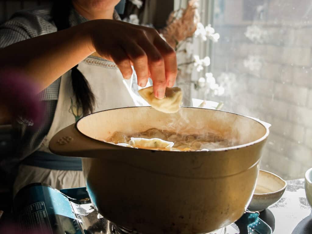 A person is cooking dumplings in a steaming pot on a stovetop. One hand is holding a dumpling above the pot. Sunlight streams through a nearby window, illuminating the kitchen area.